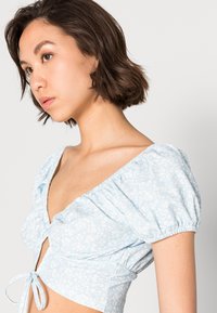 Top cropped azul claro com mangas bufantes, decote em V profundo, padrão floral e detalhe de laço na cintura. Textura de tecido suave.