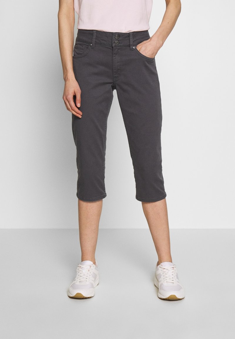 Tmavě šedé capri kalhoty s přiléhavým designem, dvěma předními kapsami a ohrnutými lemy, v kombinaci s bílými teniskami. Měkký bavlněný materiál.