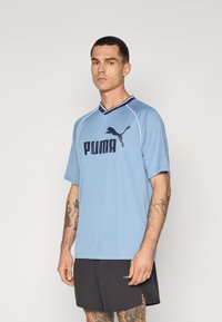 Lys blå atletisk trøje med korte ærmer, der har et sort Puma-logo og v-hals-design. Båret med sorte shorts, der viser tatoverede arme.