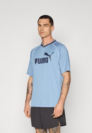 Lichtblauwe sporttrui met korte mouwen, met een zwart Puma-logo en een V-hals ontwerp. Draag met zwarte shorts, met zichtbare getatoeëerde armen.