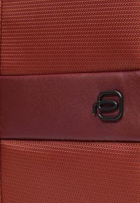 Bolsa de tela texturizada roja con un acento de cuero liso, que presenta un pequeño logotipo negro. Construcción resistente y detalles de diseño minimalistas.