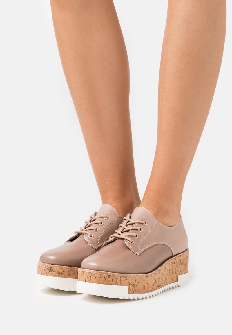 Pieds chaussés de chaussures à plateforme en cuir beige à lacets, avec des semelles compensées en liège et des semelles extérieures blanches rainurées, sur un fond uni.