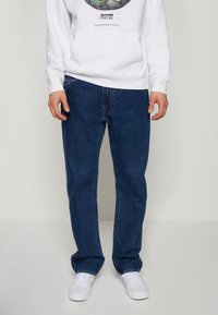 Slim-fit mörkblå jeans med urblekt textur, femfickorsdesign och bältesöglor. Bärs med en vit hoodie och vita sneakers.