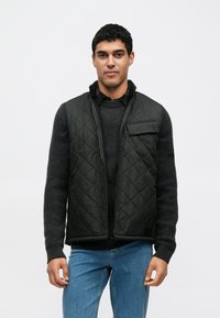 Czarna pikowana kamizelka z suwakiem z przodu, stójką i kieszenią na piersi, noszona na ciemnym teksturowanym swetrze i niebieskich dżinsach.