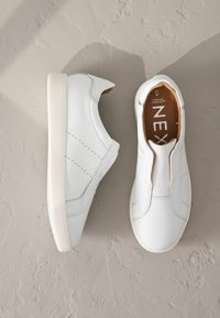 Hvide slip-on sko lavet af struktureret læder, med elastiske sideskaller, en glat rund tå og en robust hvid gummi sål.