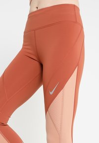 Leggings Nike a vita alta in ruggine e pesca, con accenti in rete, una trama liscia e un design curvo con logo riflettente.