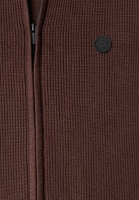 Gros plan sur un tissu tricoté côtelé marron avec une fermeture éclair noire au centre et un patch rond avec un logo noir sur le côté droit.