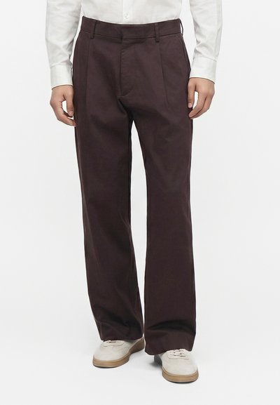 Homme portant un pantalon droit marron foncé associé à des baskets blanches et une chemise blanche à manches longues, debout devant un fond blanc.