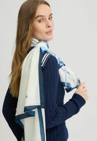 Silikone tørklæde med blomster mønster i blå nuancer, kantet med marineblå, draperet stilfuldt rundt om halsen, parret med en marineblå cable-knit sweater.
