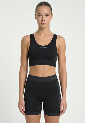 Une femme porte un soutien-gorge de sport noir Hummel et un short de cyclisme ajusté sur un fond clair uni.