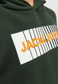 Felpa con cappuccio verde scuro caratterizzata da una grafica rettangolare bianca con strisce nere e testo arancione in grassetto che recita "JACK & JONES". Tessuto morbido e texture.