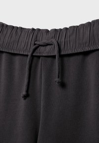 Pantaloni della tuta con elastico nero in vita e coulisse annodata al centro davanti, mostrando la trama del tessuto e i dettagli delle cuciture.