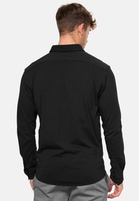 Camisa negra de manga larga con cuello, con puños abotonados y una textura suave. Diseñada con un corte ajustado y costuras en los hombros traseros.