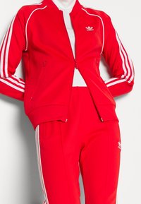 Survêtement zippé rouge en tissu lisse. Présente des accents à trois rayures blanches et un logo. La veste a des poches latérales ; le pantalon a une taille élastique.