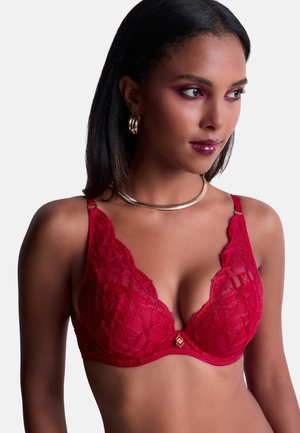 Femme portant un soutien-gorge en dentelle rouge avec des boucles d'oreilles et un collier dorés, regardant vers la droite avec un maquillage subtil et des cheveux lisses et foncés.