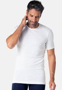 T-shirt blanc à manches courtes en tissu lisse et extensible ; col rond ; porté par un homme barbu, se tenant devant un fond neutre.