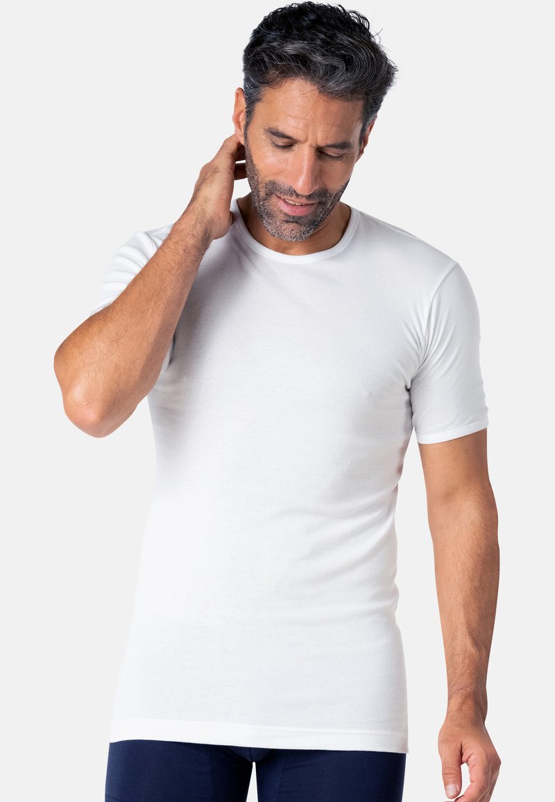 T-shirt blanc à manches courtes en tissu lisse et extensible ; col rond ; porté par un homme barbu, se tenant devant un fond neutre.