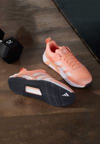Light peach athletic shoes with white accents, textured mesh upper, and a black rubber sole featuring a tread pattern, resting on wooden surface.