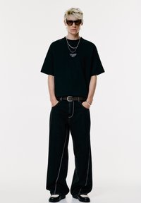 Giovane con capelli biondi corti che indossa occhiali da sole scuri, una maglietta nera oversize con testo, jeans neri larghi e scarpe in bianco e nero.