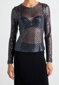 Žena v černém průhledném topu s dlouhým rukávem a stříbrným flitrovým diamantovým vzorem přes černou podprsenku a sukni.