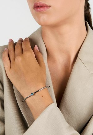 Bracciale rigido in argento con un accento di pietra blu, indossato su una mano appoggiata su un tessuto di tonalità neutra. Finitura metallica liscia.