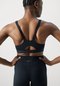 Svart sports-bh med brede stropper, utskåret rygg, justerbare kroker og preget "NIKE"-logo. Kombinert med svarte høyt-hengende leggings.