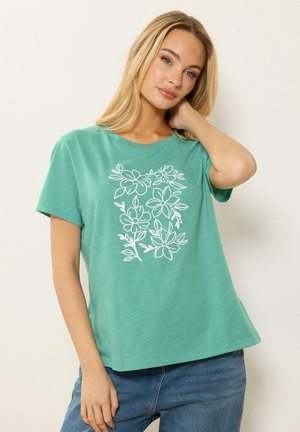 Nuori nainen, jolla on pitkät vaaleat hiukset ja joka pukeutuu petroolinsiniseen lyhythihaiseen paitaan, jossa on valkoinen kukkakuvio, sekä sinisiin farkkuihin, poseeraa käsi kaulansa lähellä.