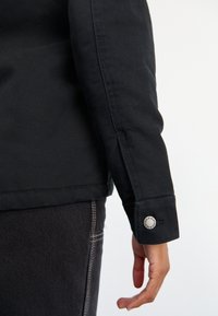 Personas labā roka un plauksta, kas ģērbušies melnā garā piedurknē t-kreklā ar Dickies metāla pogu un tumšajos džinsos.