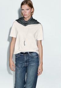Witte katoenen T-shirt gecombineerd met blauwe denim jeans. Een grijze sjaal hangt over de schouders. Eenvoudige zilveren hanger en een zwarte riem.