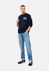 Felpa navy con logo, jeans azzurri chiaro e sneaker scure. Vestibilità casual, tessuto testurizzato, scollatura semplice e maniche arrotolate.