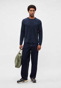 Tumši zils krekls ar garām piedurknēm un kontrastējošām šuvēm, kopā ar vaļīgām tumša džinsa biksēm un daudzkrāsainām sporta kurpēm. Rokā tiek turēta olīvu zaļa soma.