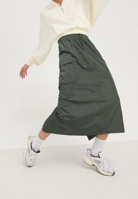 Gonna midi verde con vita regolabile e tasche laterali, abbinata a un pullover crema e scarpe da ginnastica grigie su uno sfondo neutro.