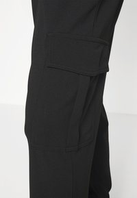 Gros plan sur un pantalon noir avec une grande poche latérale à rabat au niveau de la cuisse.