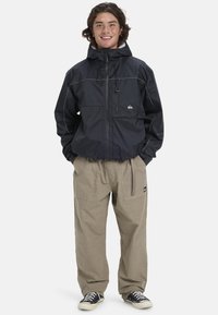 Black waterproof jacket with adjustable hood, front zip, and chest pocket. Paired with light beige cargo pants and black sneakers.