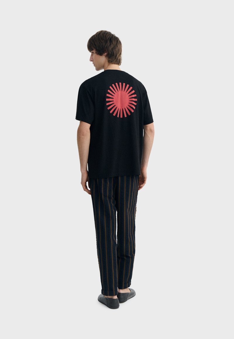 Jeune homme vu de dos portant un t-shirt noir avec un grand motif de rayon de soleil rouge et un pantalon rayé sombre avec des chaussures noires.
