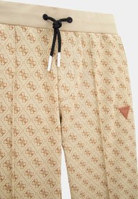 Beiga joggers med diamantmönster och logotyp, med svart dragsnöre och ribbad midja, samt en brun etikett på sidan.