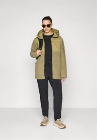 Kvinne iført solbriller, olivengrønn quilted og fleecejakke, svarte bukser, beige sneakers, bærende på en svart ryggsekk, stående mot en ensfarget bakgrunn.