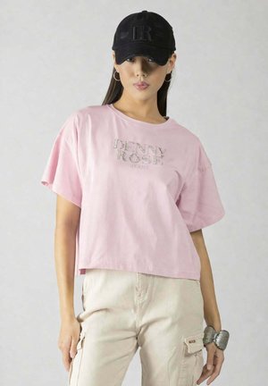 Donna che indossa una t-shirt rosa "Denny Rose", pantaloni cargo beige, cappellino nero con logo "DR", orecchini a cerchio in argento e un grande bracciale in argento.