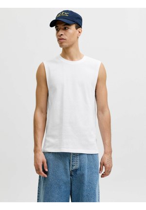 Mees, kes kannab valget varrukateta särki, lahtisi siniseid pükse ja tumelilla nokamütsi, seisab ühevärvilise heleda tausta ees.