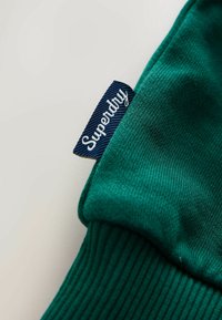 Zielona bluza z dzianiny z prążkowanymi mankietami, z granatową metką z napisem "Superdry" w białym kolorze. Gładka tekstura i casualowy design.