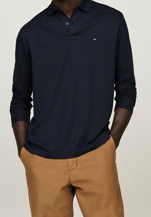 Homme portant un polo bleu marine à manches longues avec un petit logo sur la poitrine et un pantalon marron clair, mains dans les poches, sur fond uni.