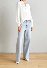 Kvinde iført en hvid langærmet bluse med flæser ved ærmerne og lyseblå høj talje-bredbenede jeans, stående på trægulv.