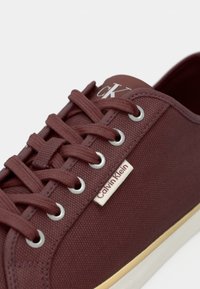 Bordeau platneni tenisice s metalnim okovima, okruglim vezicama i oznakom Calvin Klein sa strane. Imaju bijeli gumeni potplat.