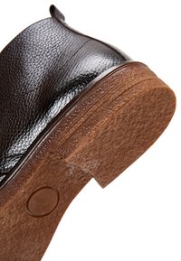 Chaussure habillée en cuir noir montrant une semelle en caoutchouc brun texturée avec un petit logo circulaire, vue rapprochée en angle.