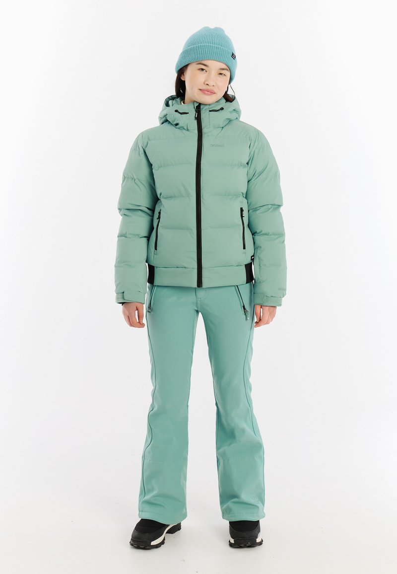 Helle grüne Steppjacke mit Kapuze, schwarzem Reißverschluss und zwei seitlichen Taschen, kombiniert mit passenden, schmal geschnittenen Schneehosen und schwarzen Schneestiefeln.