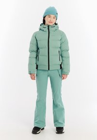 Lichtgroene pufferjack met capuchon, zwarte rits en twee zijzakken, gecombineerd met bijpassende slim-fit sneeuwbroeken en zwarte sneeuwlaarzen.