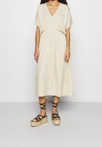 Frau in einem beigen Midikleid mit weißen Punkten und schwarzen Plateau-Sandalen mit Knöchelriemen vor einem einfarbigen Hintergrund.