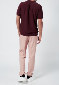 Mies, joka on pukeutunut viininpunaisiin lyhythihaisiin paitoihin, vaaleanpunaiseen housuihin ja valkoisiin tennareihin, joissa on mustat kantakohdat, seisoo selkä kameraan päin.