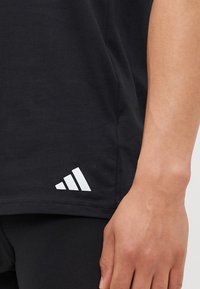 Fekete pamut T-shirt kerek nyakkivágással, fehér Adidas logóval és három átlós csíkkal az alján. Közeli kép az ujjon és az oldalon.