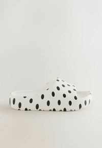 Sandale blanche à enfiler avec des pois noirs, bout ouvert, semelle épaisse et dessous texturé pour une meilleure adhérence, vue de profil sur un fond neutre.